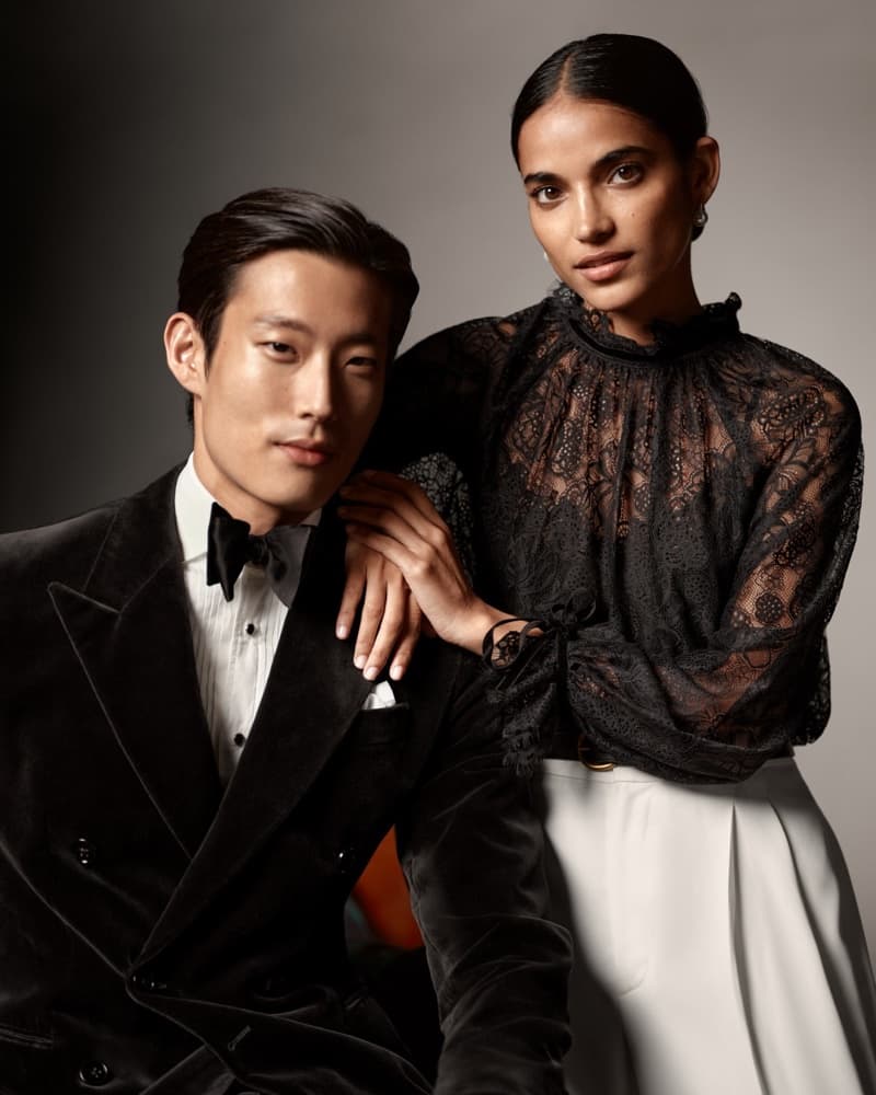 An Asian man in a black tuxedo and a Hispanic woman in a black lace blouse pose together in a medium shot against a gray backdrop.