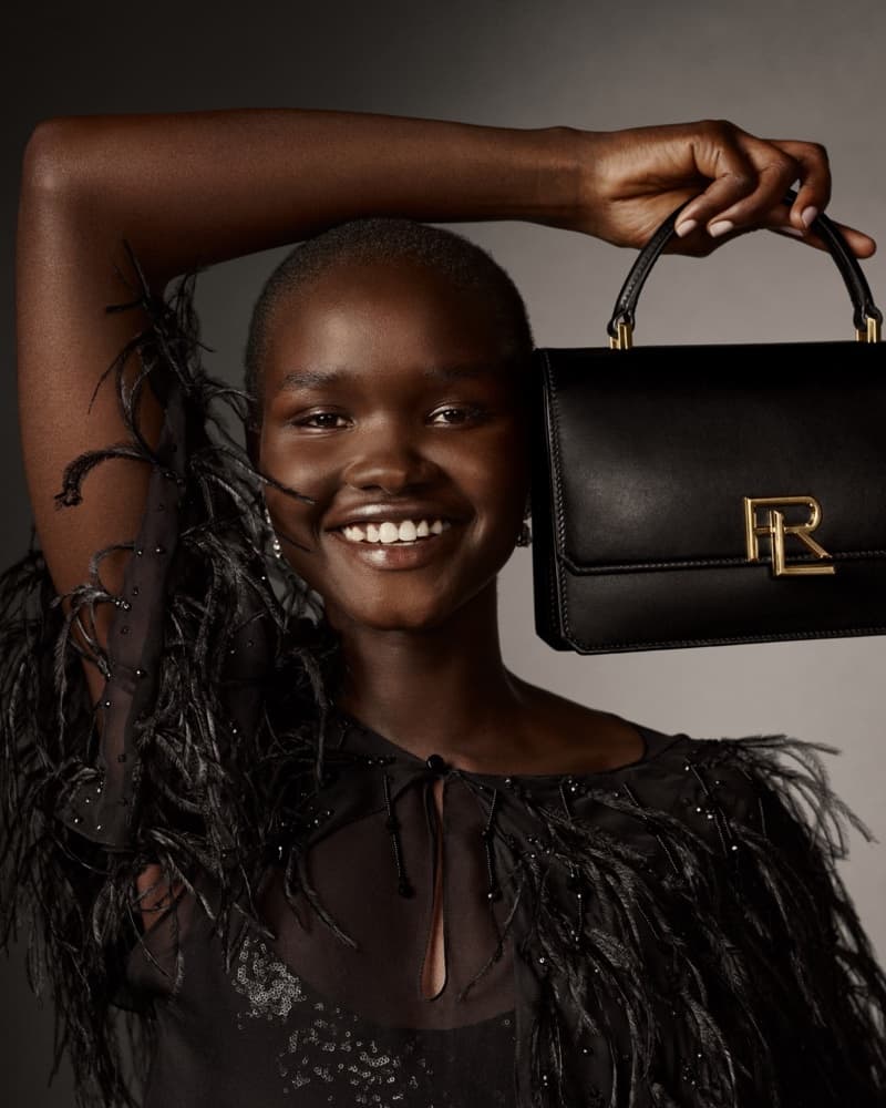 Smiling adult woman in a feathered top is holding a black leather handbag with a gold-colored logo against a gray background.
