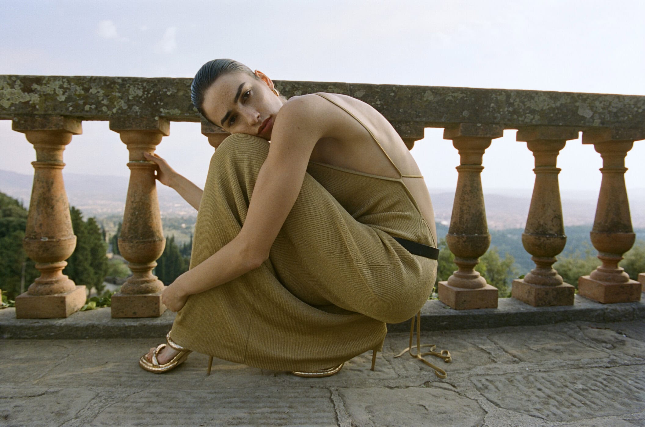 Woman in flowing gold pants and tank top posing against a weathered stone railing with a scenic green landscape backdrop on a sunny day.