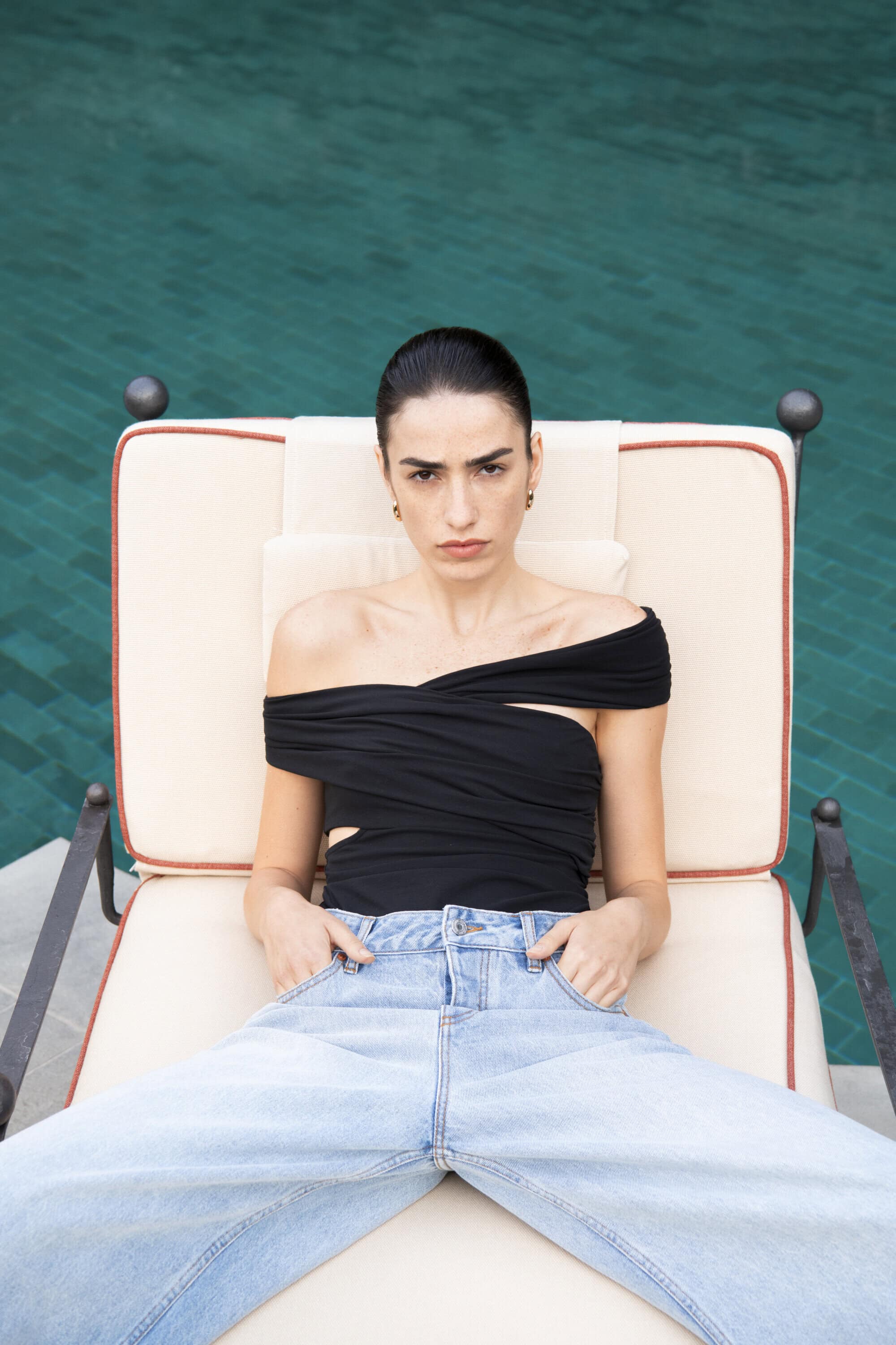Adult female model poses on a lounge chair by a pool, wearing a black top and light-wash denim jeans in an outdoor setting.