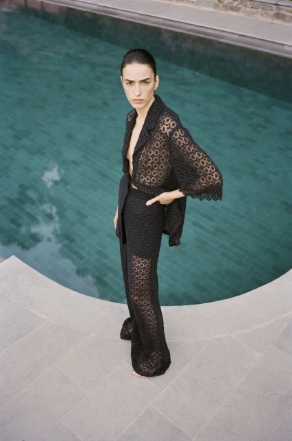 Woman in a sheer black lace-patterned two-piece suit poses by a pool with turquoise water and gray stone tiling on the deck.