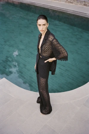 Woman in a sheer black lace-patterned two-piece suit poses by a pool with turquoise water and gray stone tiling on the deck.