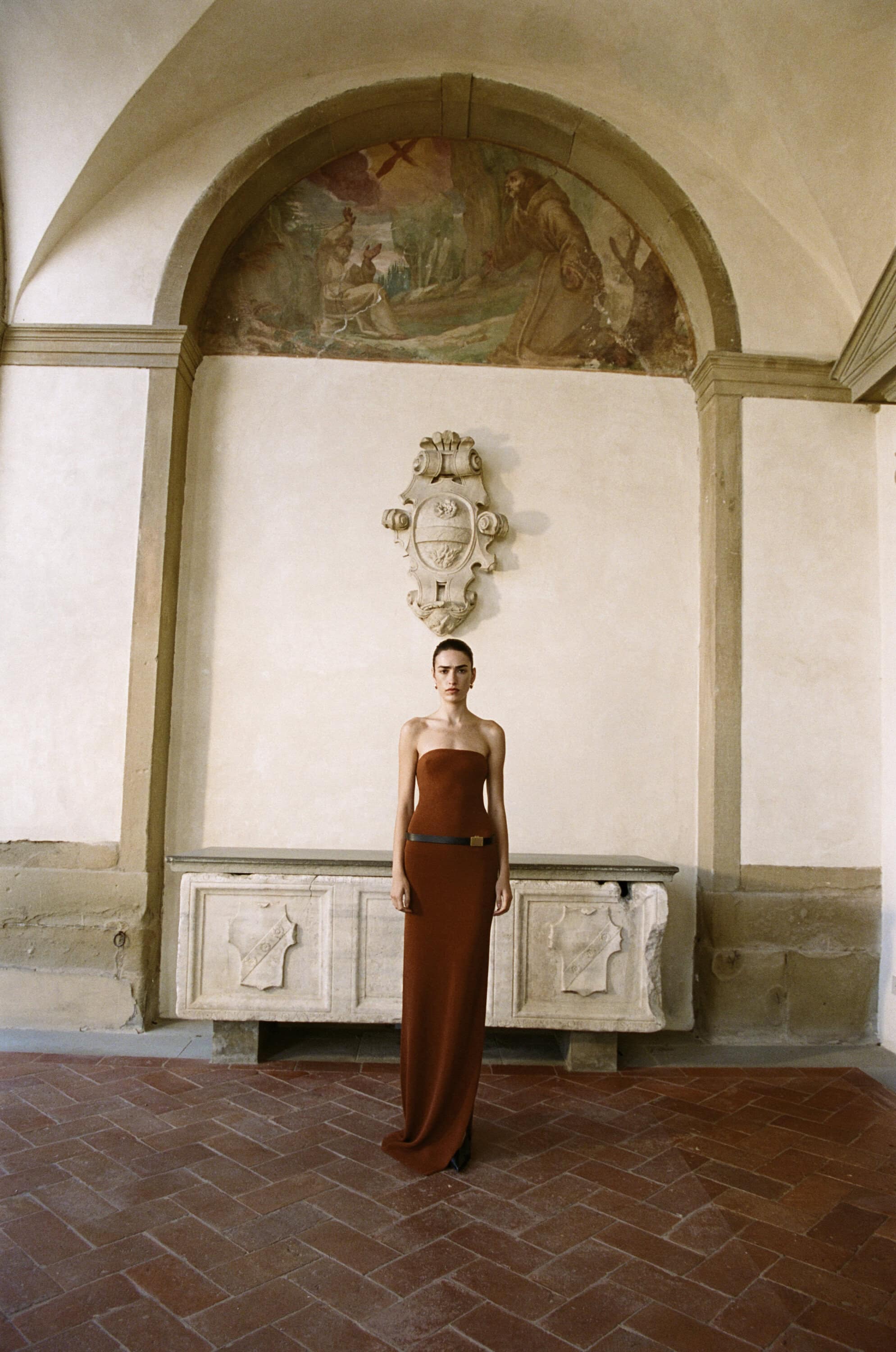 A slender woman stands in a strapless, floor-length gown in a spacious interior with high arches and a Renaissance mural.