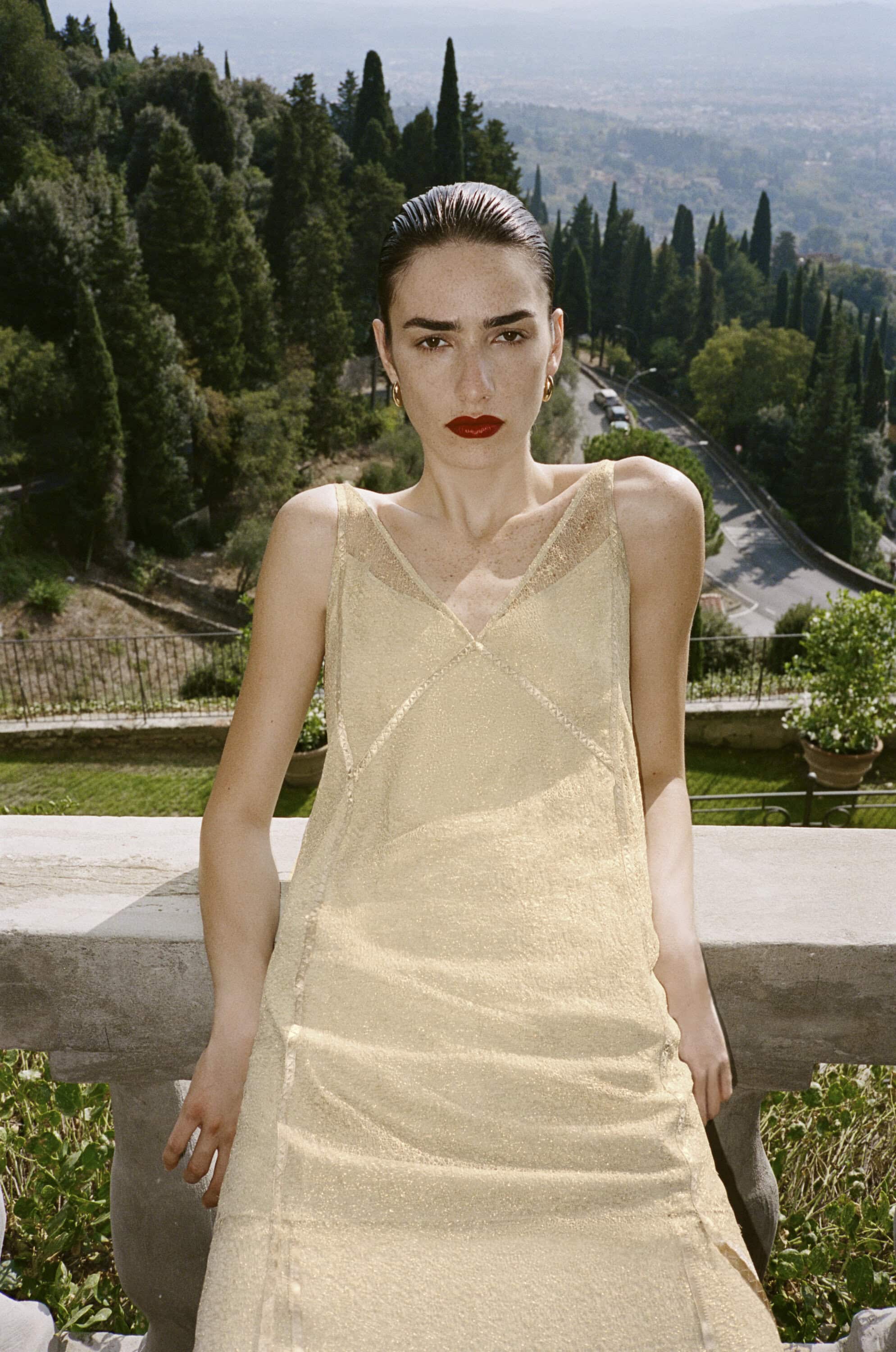 A stylish woman in a champagne-colored dress stands on a stone balcony with a sprawling Tuscan landscape in the background, fashion shoot.