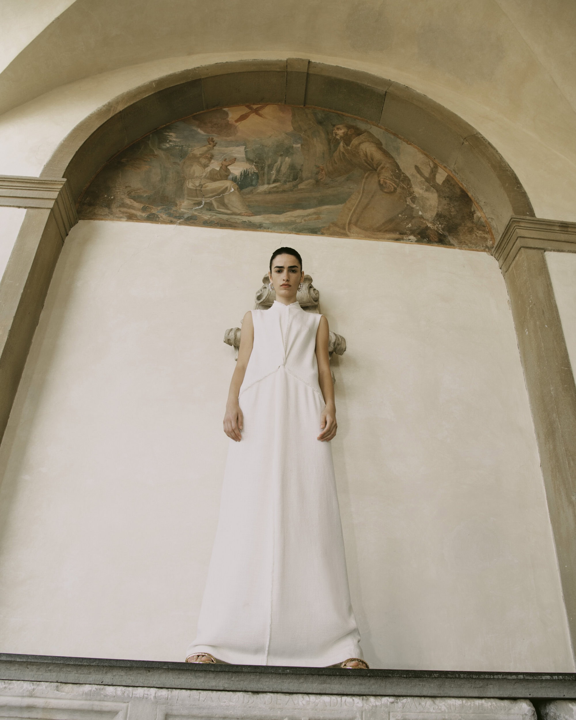 A woman in a white dress stands in front of a wall with a fresco. The light is soft, and the architectural setting is historic.