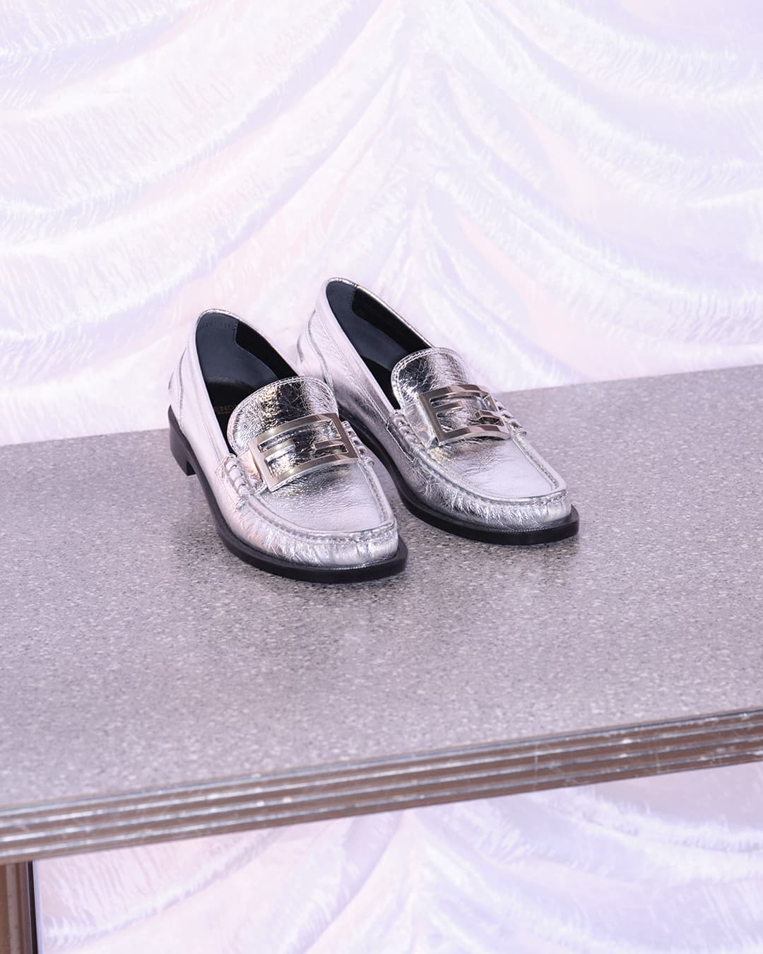 Eye-level studio shot of silver penny loafer shoes on a metallic surface with a white textured backdrop, showcasing fashion footwear.