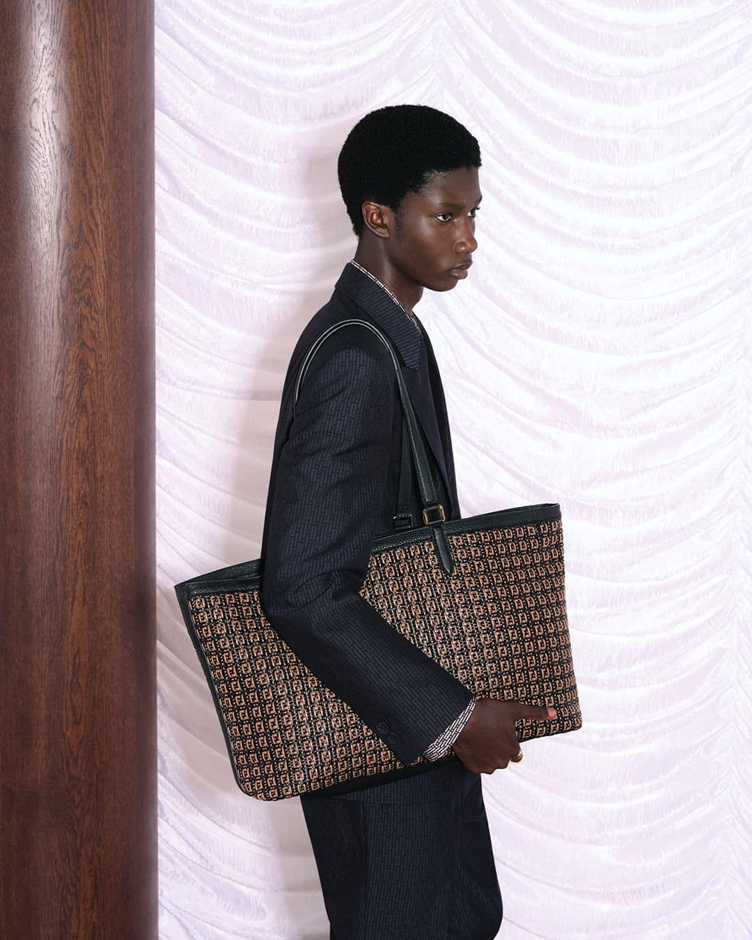 A dark-skinned man in a suit carries a designer tote bag in an indoor setting with soft lighting, showcasing fashion and style.