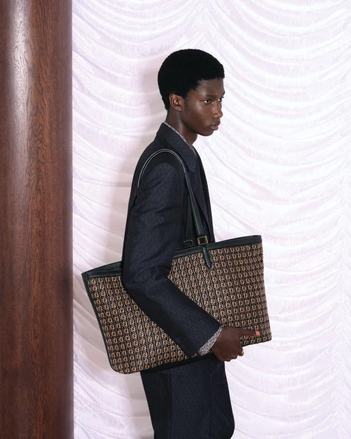 A dark-skinned man in a suit carries a designer tote bag in an indoor setting with soft lighting, showcasing fashion and style.