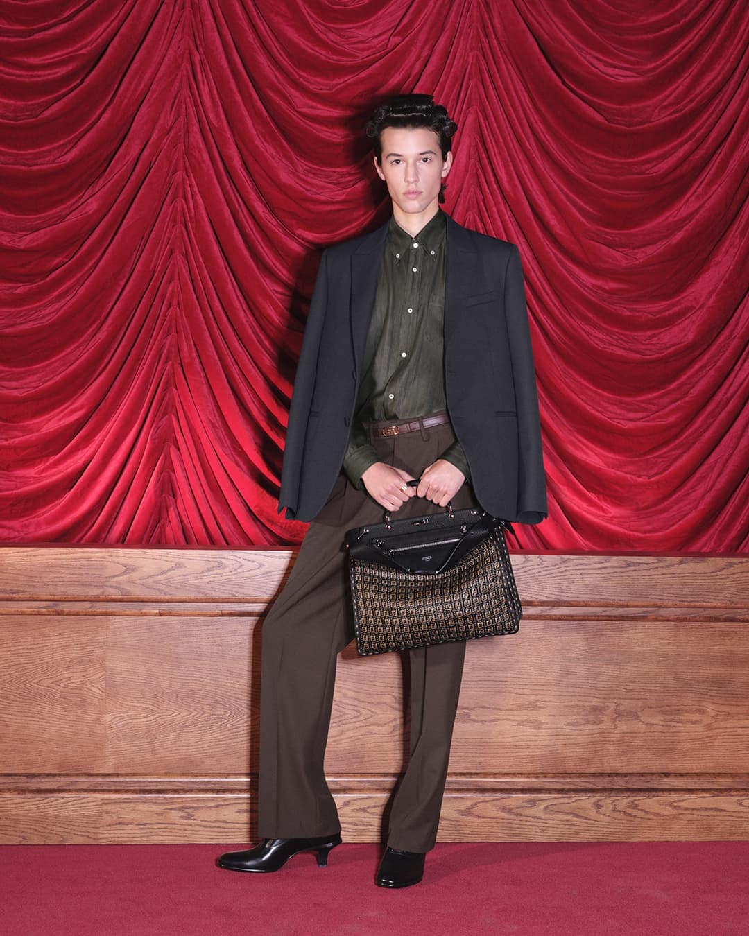 Male model wearing a suit and holding a handbag, posing in front of a red curtain and wood paneling in an indoor setting.