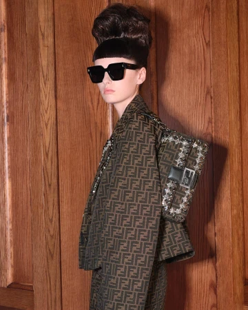 Brunette woman in patterned Fendi suit and sunglasses poses in front of wood wall with a Fendi handbag on her shoulder in a studio shot.