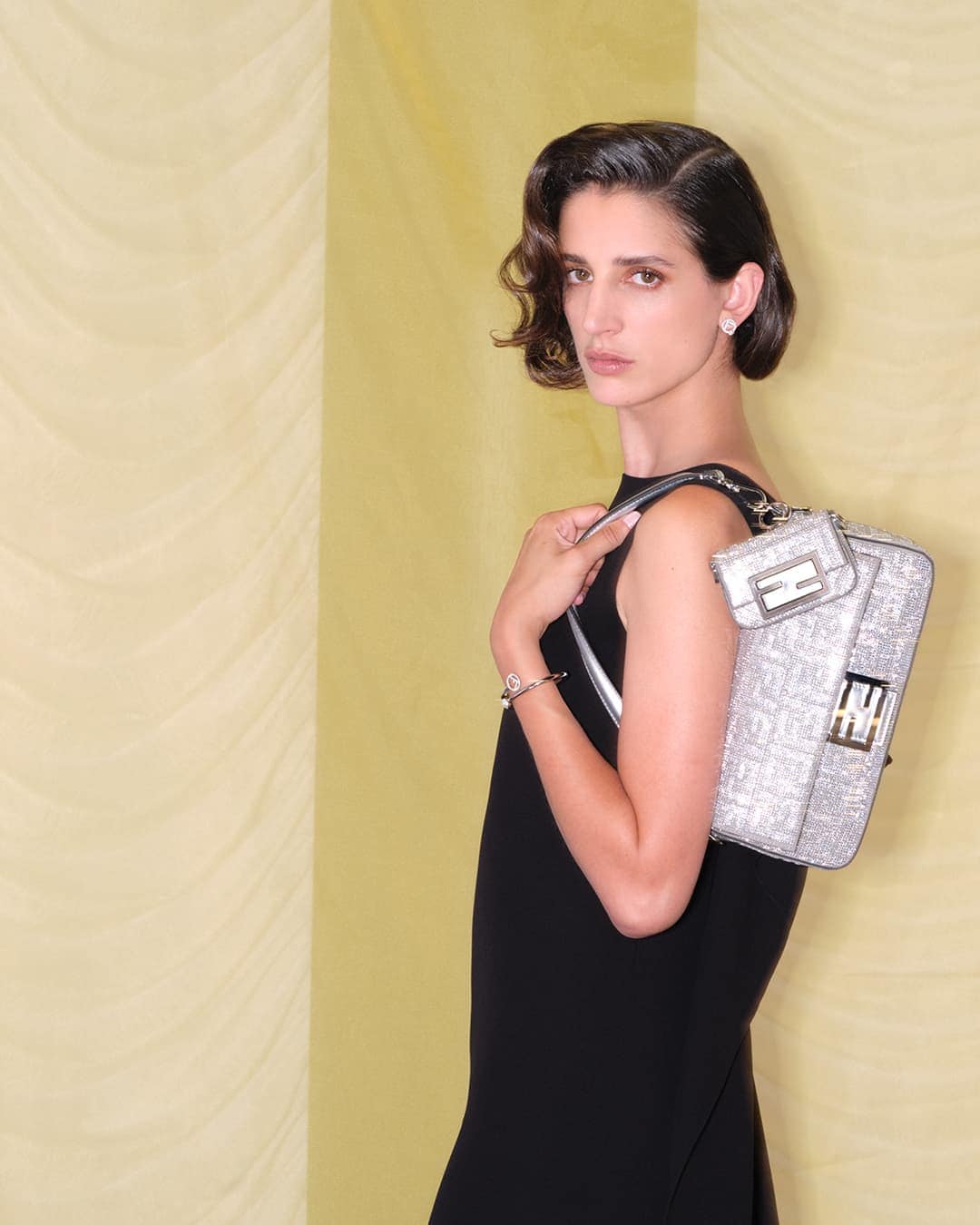 Adult woman in black dress poses with silver Fendi handbag in a studio with tan and yellow striped background for a fashion shot.