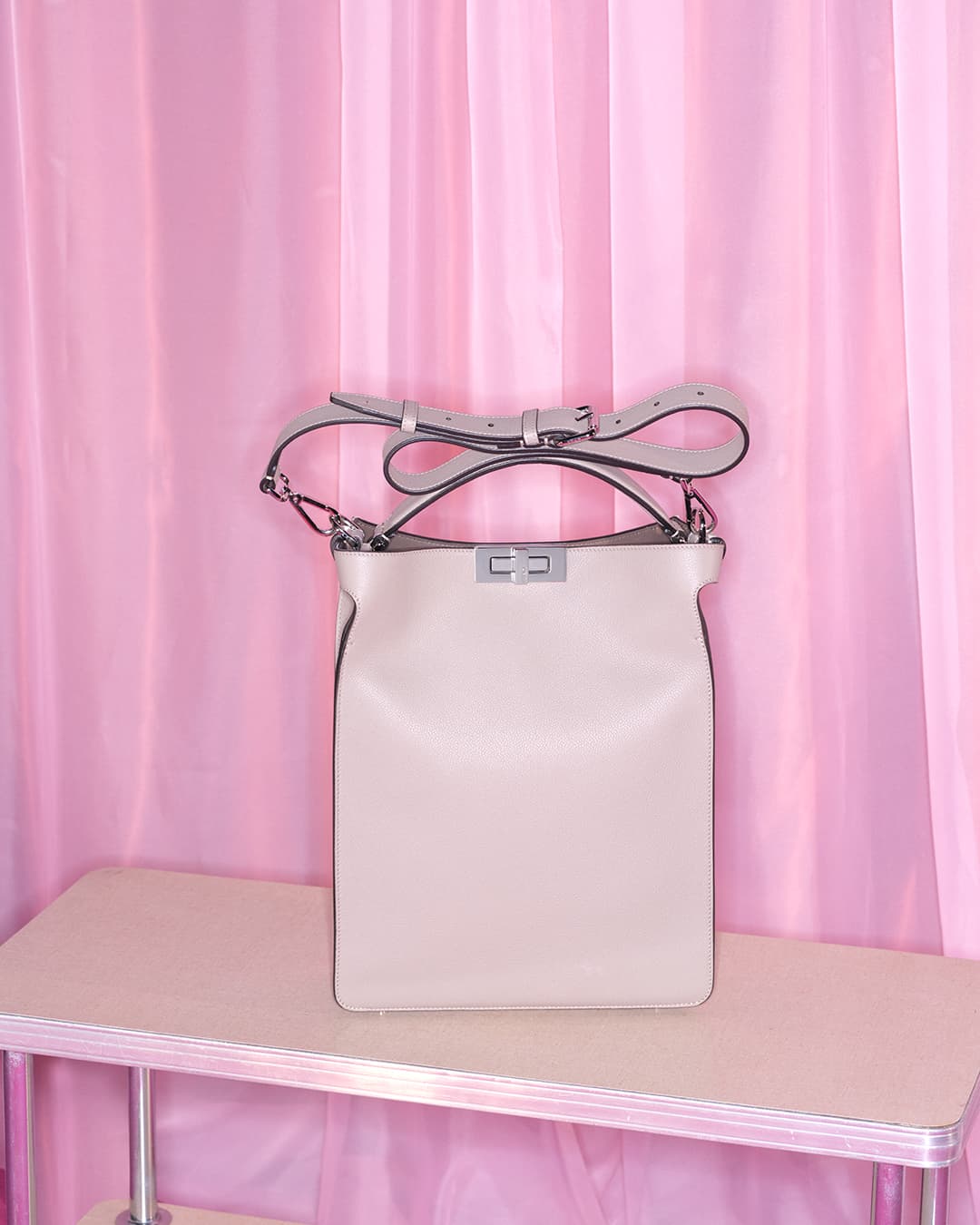 Studio shot of a light beige leather satchel handbag with silver hardware, displayed on a beige table against a pink curtain backdrop.
