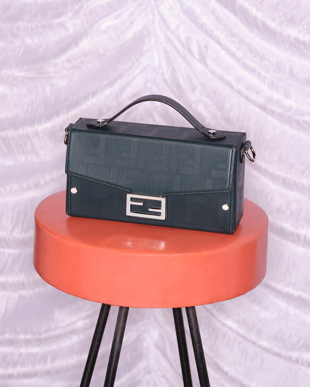 Green textured Fendi handbag on a coral-colored stool with black metal legs, set against a backdrop of white patterned fabric.
