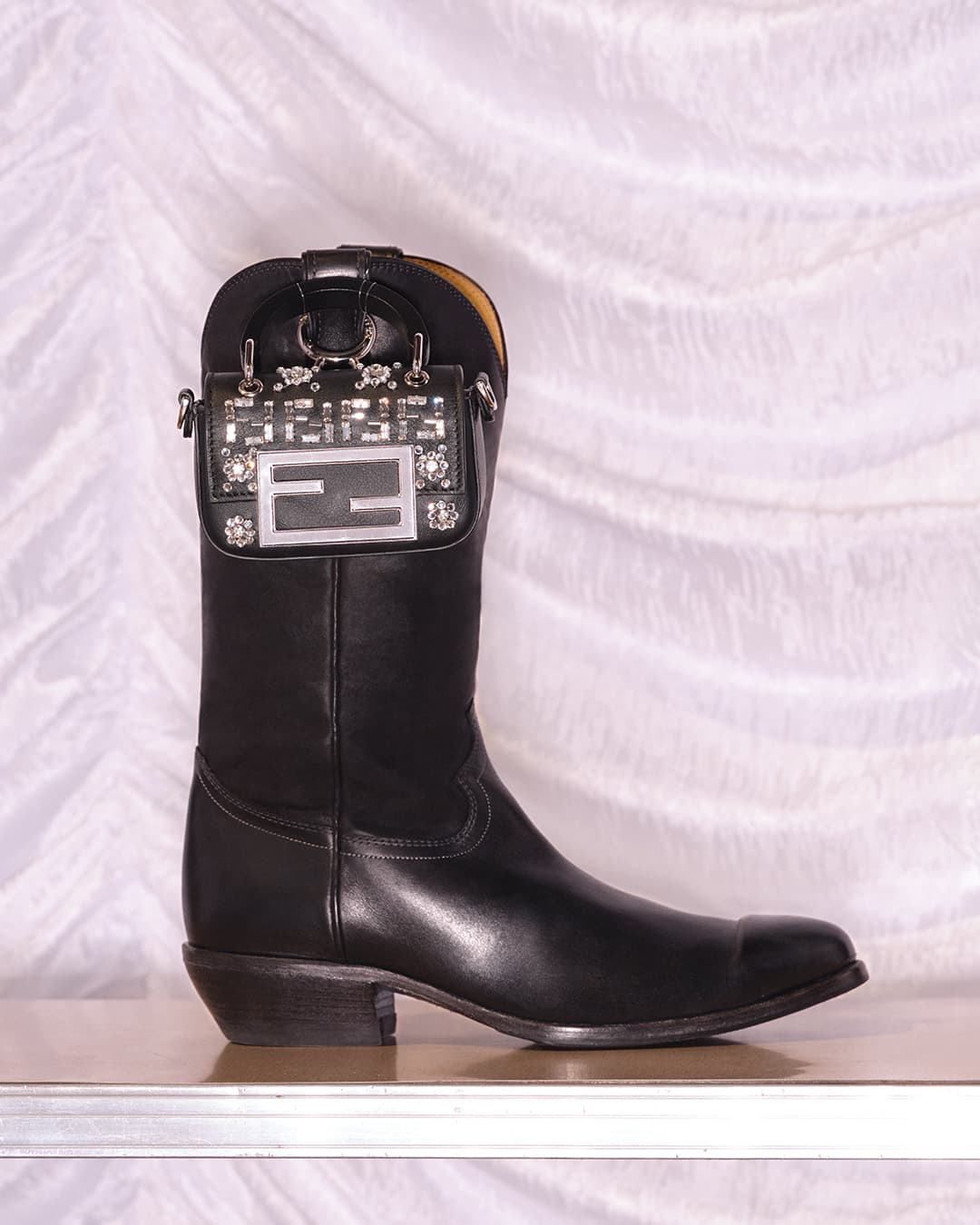 Leather cowboy boot with embellished Fendi mini bag attached, against a white textured backdrop. Fashion still life photography.