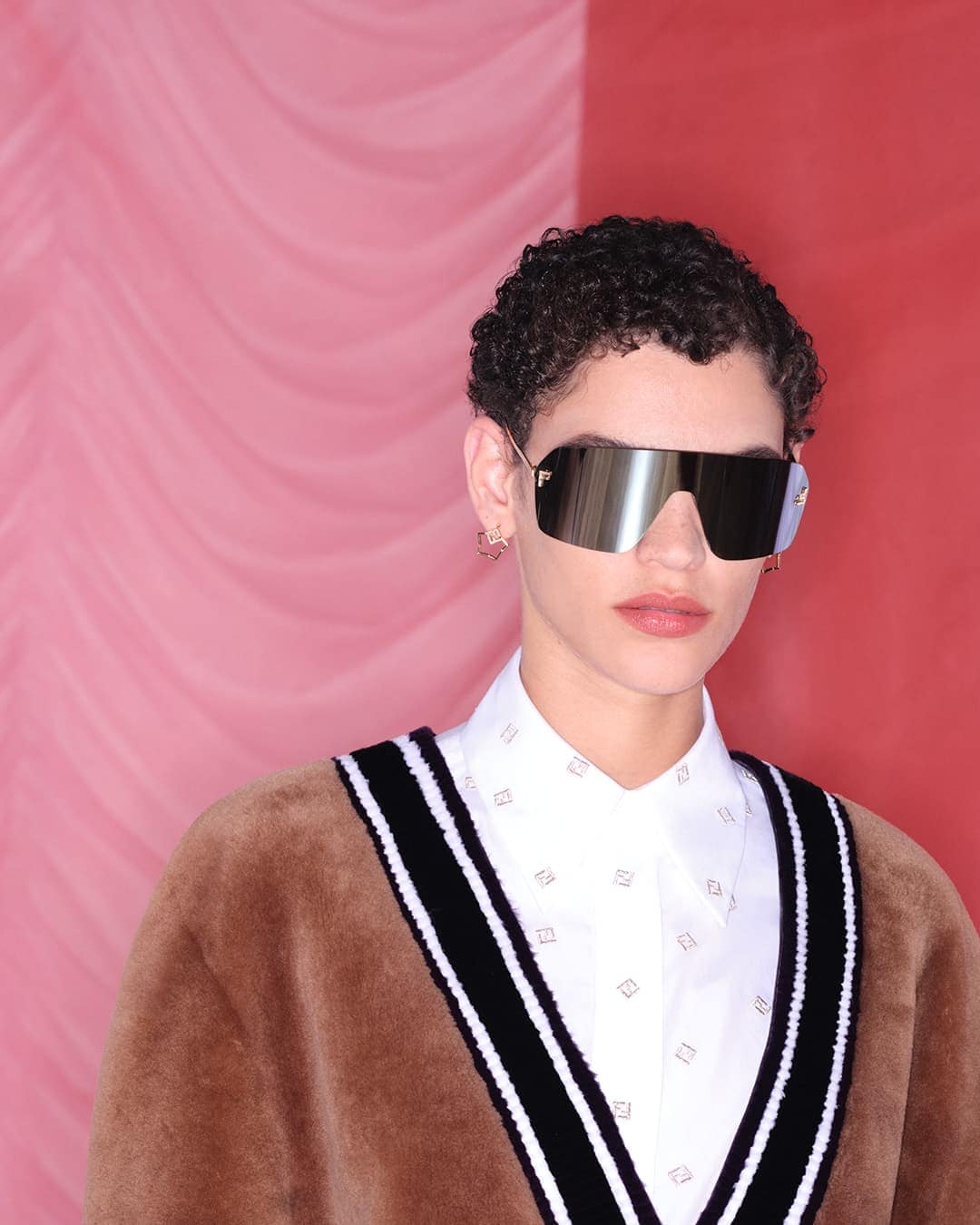 A young adult woman poses in a brown sweater and silver sunglasses against a backdrop with vertical pink and red color blocks.
