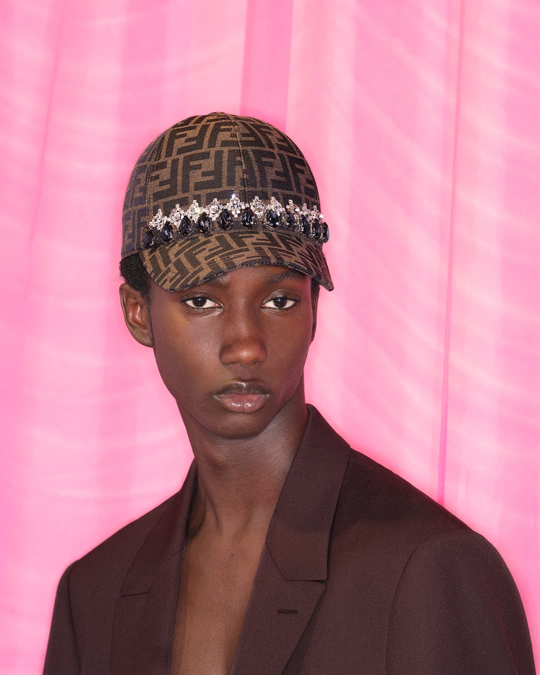 Young man in a patterned ball cap with jeweled trim poses against a vibrant pink backdrop, showcasing a modern fashion statement.