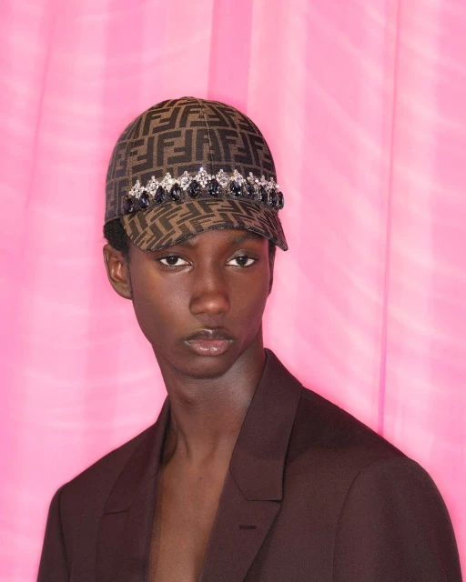Young man in a patterned ball cap with jeweled trim poses against a vibrant pink backdrop, showcasing a modern fashion statement.