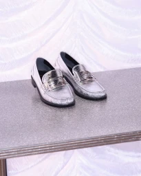 Pair of metallic silver leather loafers with logo buckles displayed on a grey textured tabletop against draped white fabric.