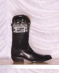 Black leather cowboy boot adorned with a miniature embellished logo bag against a draped white fabric background.