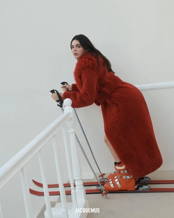 A woman in a red coat and ski boots poses on skis on an indoor staircase, looking back at the camera. Neutral, well-lit background.