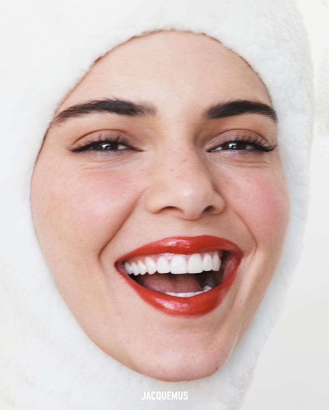 Close-up portrait of a smiling woman with bright red lipstick wearing a fluffy white hood, captured in bright, even lighting.
