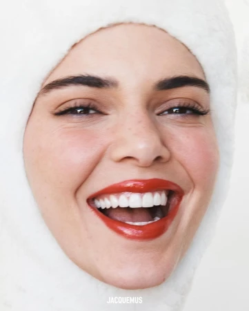 Close-up portrait of a smiling woman with bright red lipstick wearing a fluffy white hood, captured in bright, even lighting.