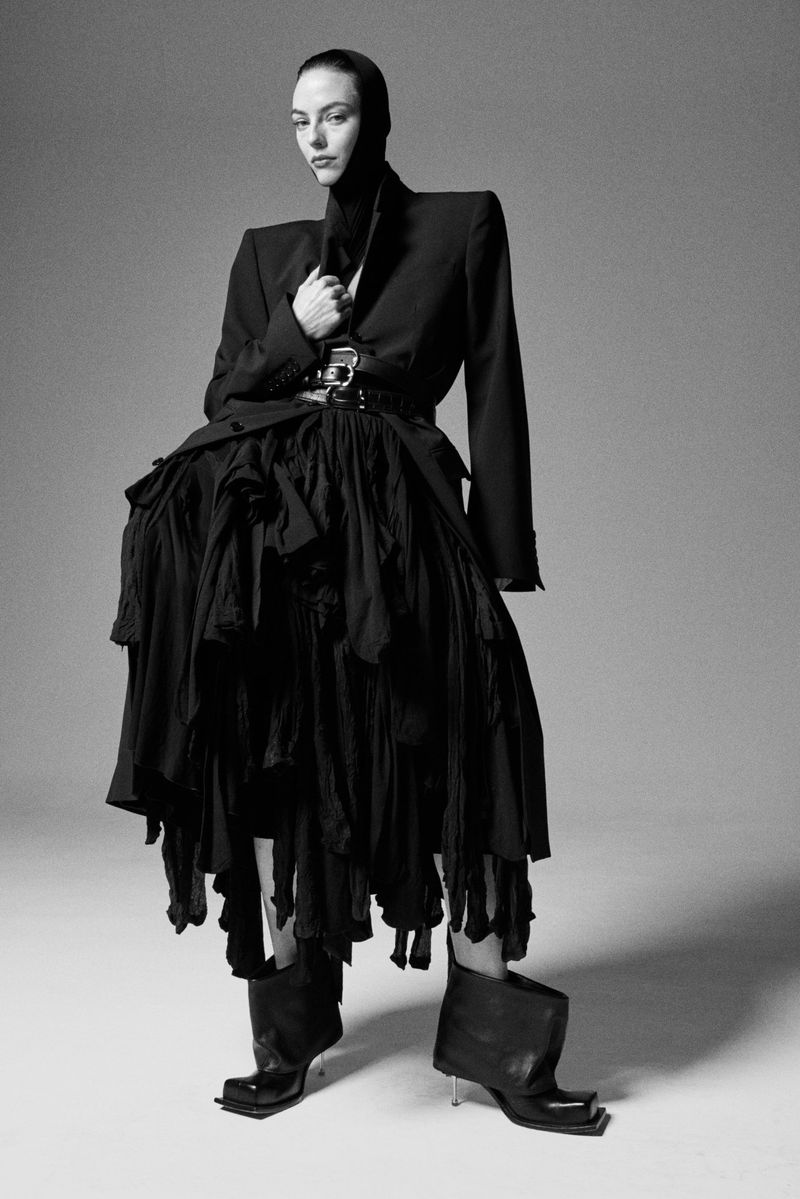 Studio shot of a fashion model in a black blazer, skirt, and headdress, posing against a gray backdrop with dramatic lighting.