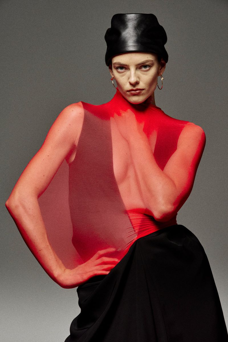 Fashionable woman poses in red sheer top, black leather headpiece, and a black skirt against a neutral gray backdrop.