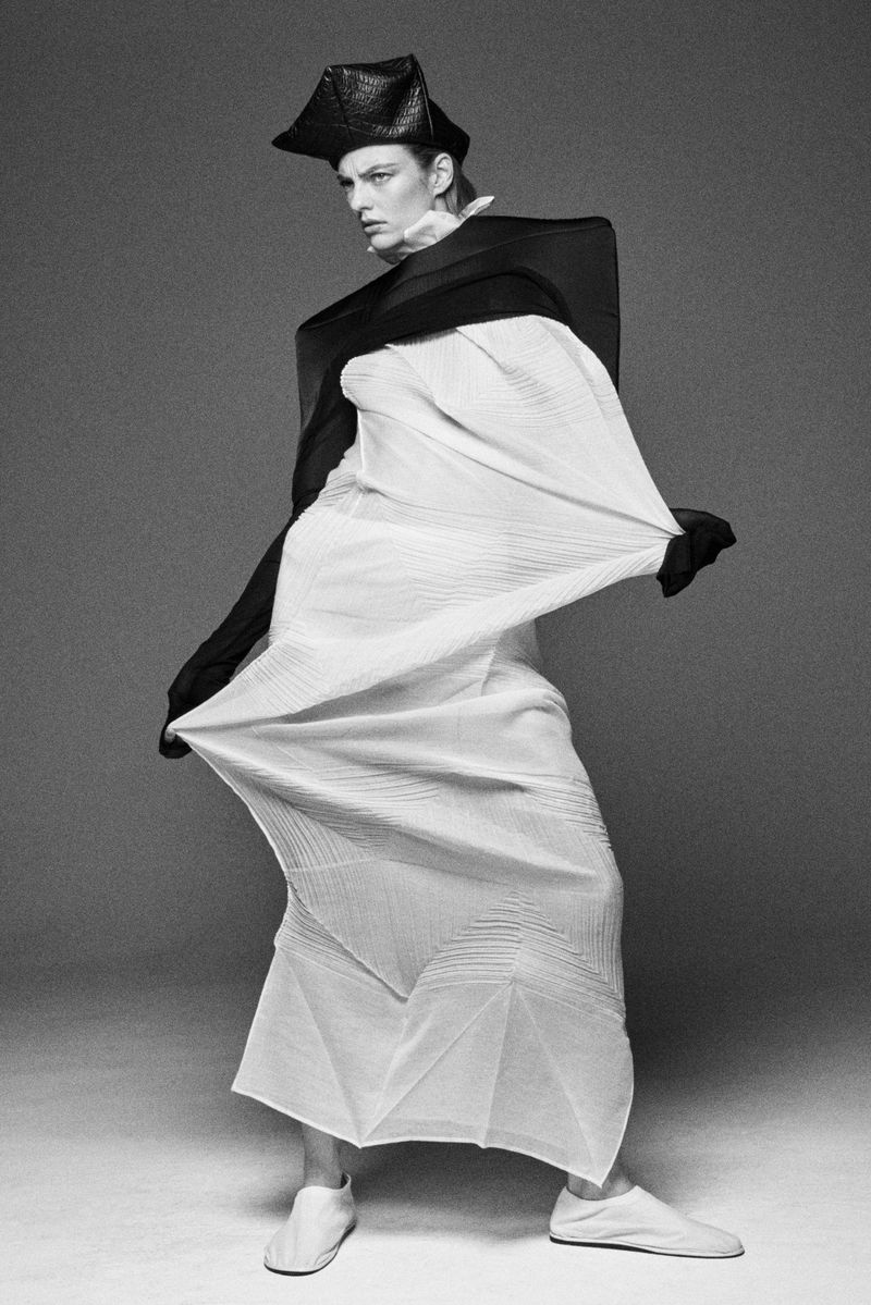 Monochrome fashion shot of a slender female model in an origami-style white dress and black hat, standing against a gray background.