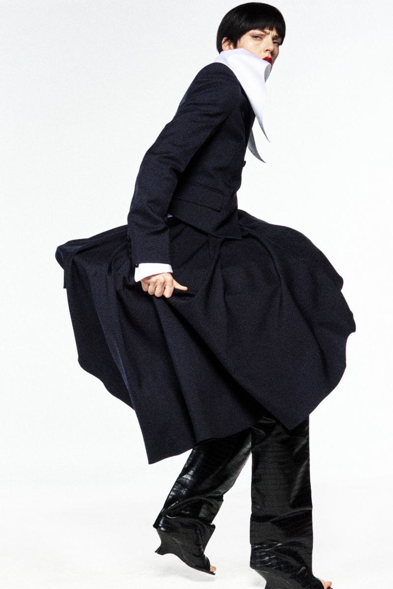 Fashion model in motion wearing a navy blazer and full skirt, paired with patent leather pants and unique open-toe heels on a white background.
