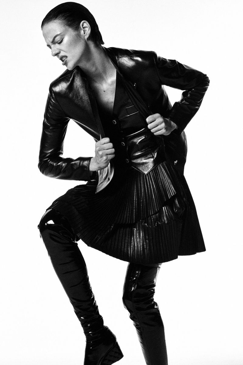 A stylish woman in a black leather jacket, vest, and skirt with thigh-high boots poses against a white background in a studio shot.