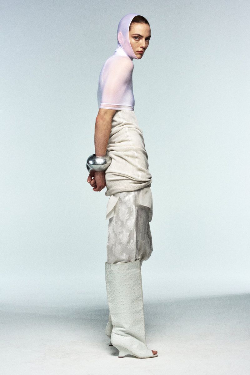 A fashion shot of a slender woman with a sheer top, draped skirt, textured leggings, and metallic bangle bracelets in a studio setting.