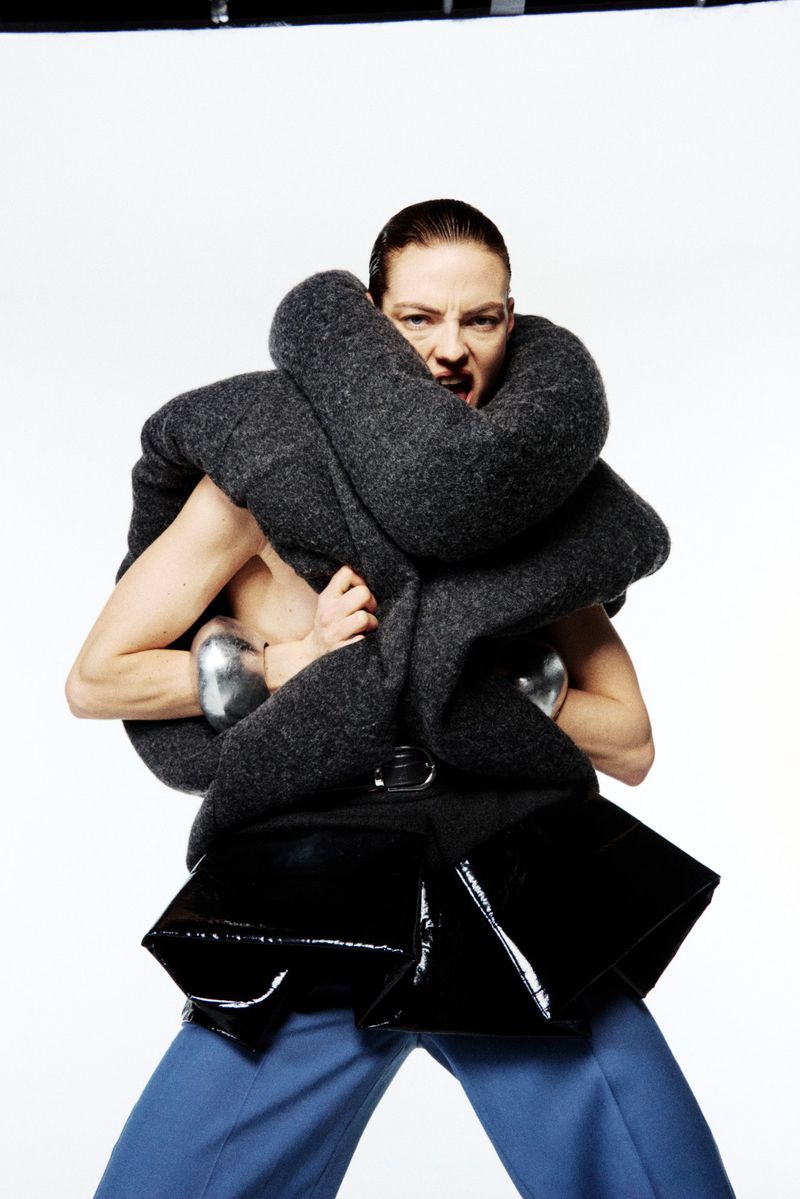 Adult female wearing an oversized grey wool scarf, shiny silver arm cuffs, a black belt with a patent leather peplum and blue trousers.