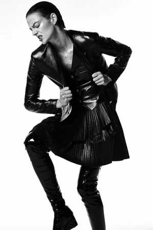 Model in black leather jacket and pleated skirt wearing high boots against a plain background