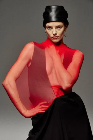 Model wearing a sheer red top and dark sculptural hat posing against a plain grey studio background.