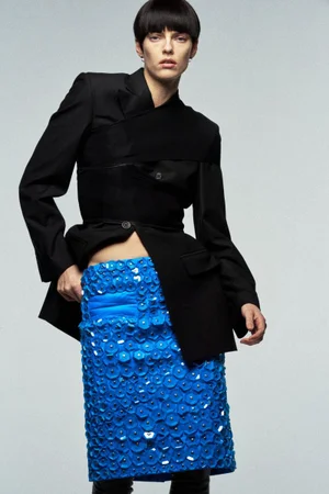 Model with a bowl cut wearing a structured black blazer and textured bright blue skirt against a pale grey studio background.