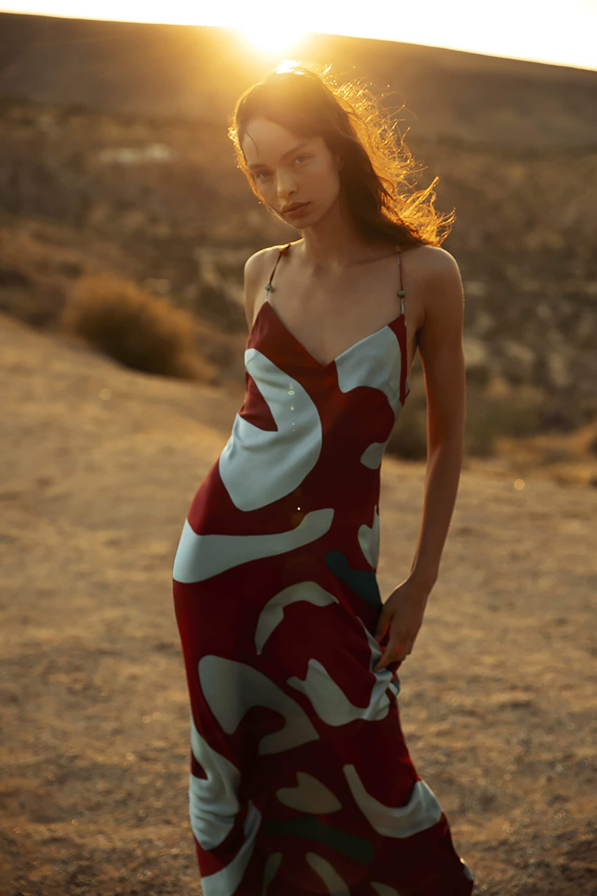 Woman in a patterned dress standing on a desert landscape at sunset. Fashion shot with warm lighting and natural setting.