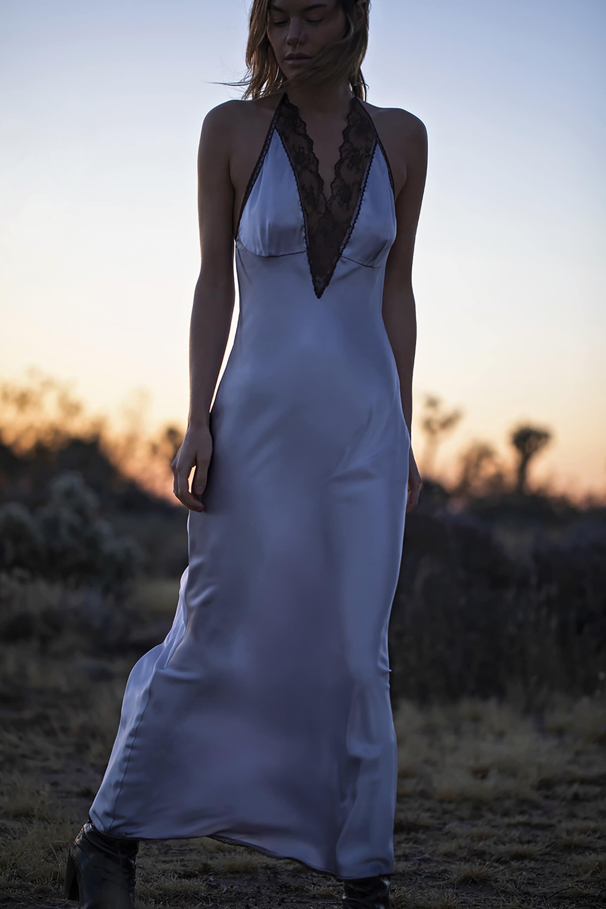 A woman stands in a desert landscape at dusk wearing a pale blue floor-length silk dress with a dark lace-trimmed halter neckline.