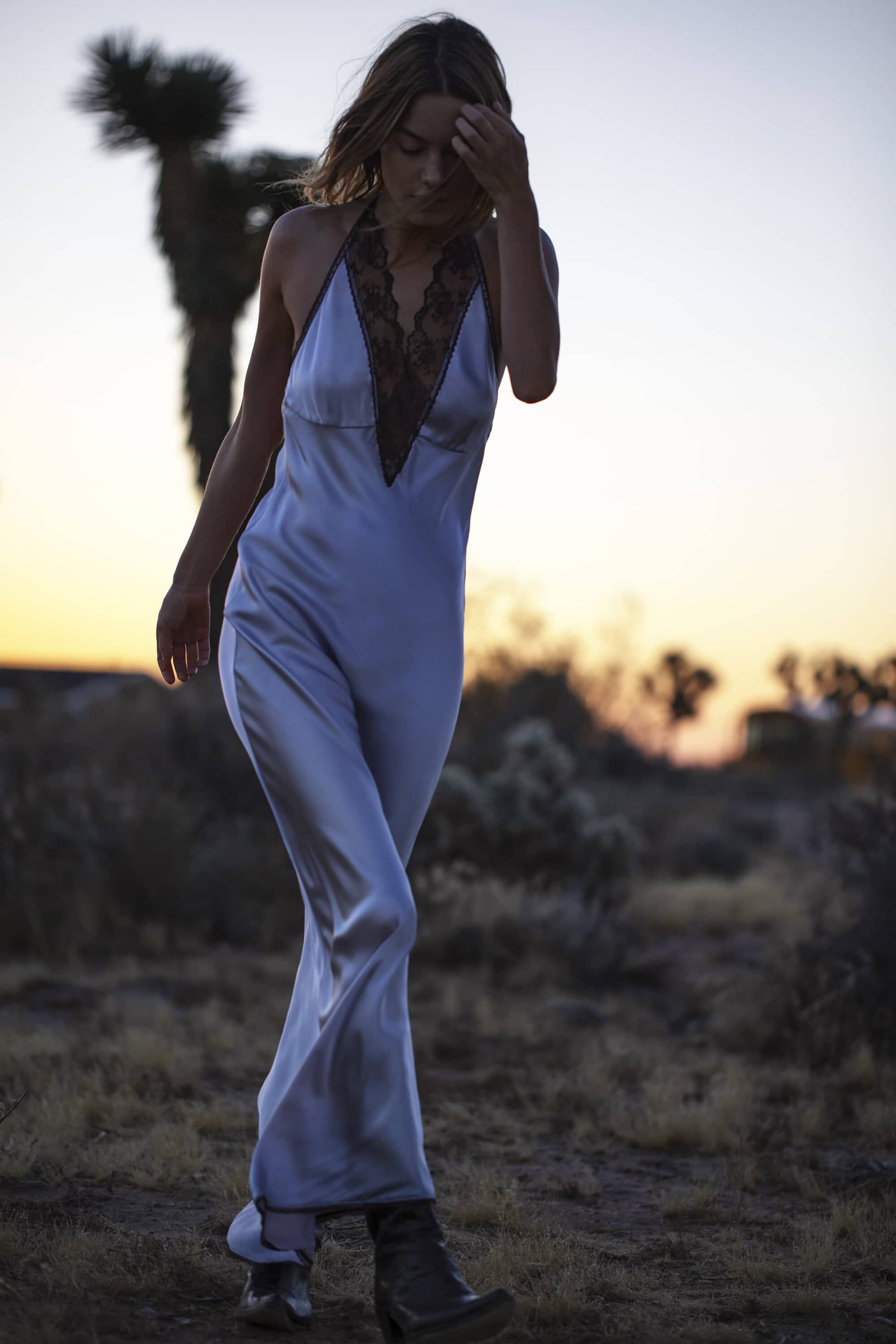 A woman in a light blue dress walks through the desert landscape at sunset, a Joshua tree visible in the background.