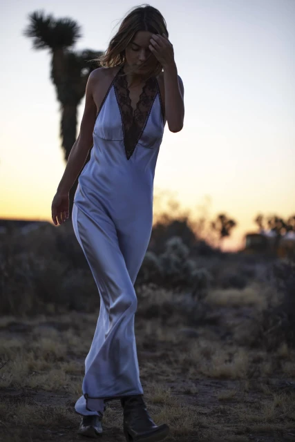A woman in a light blue dress walks through the desert landscape at sunset, a Joshua tree visible in the background.