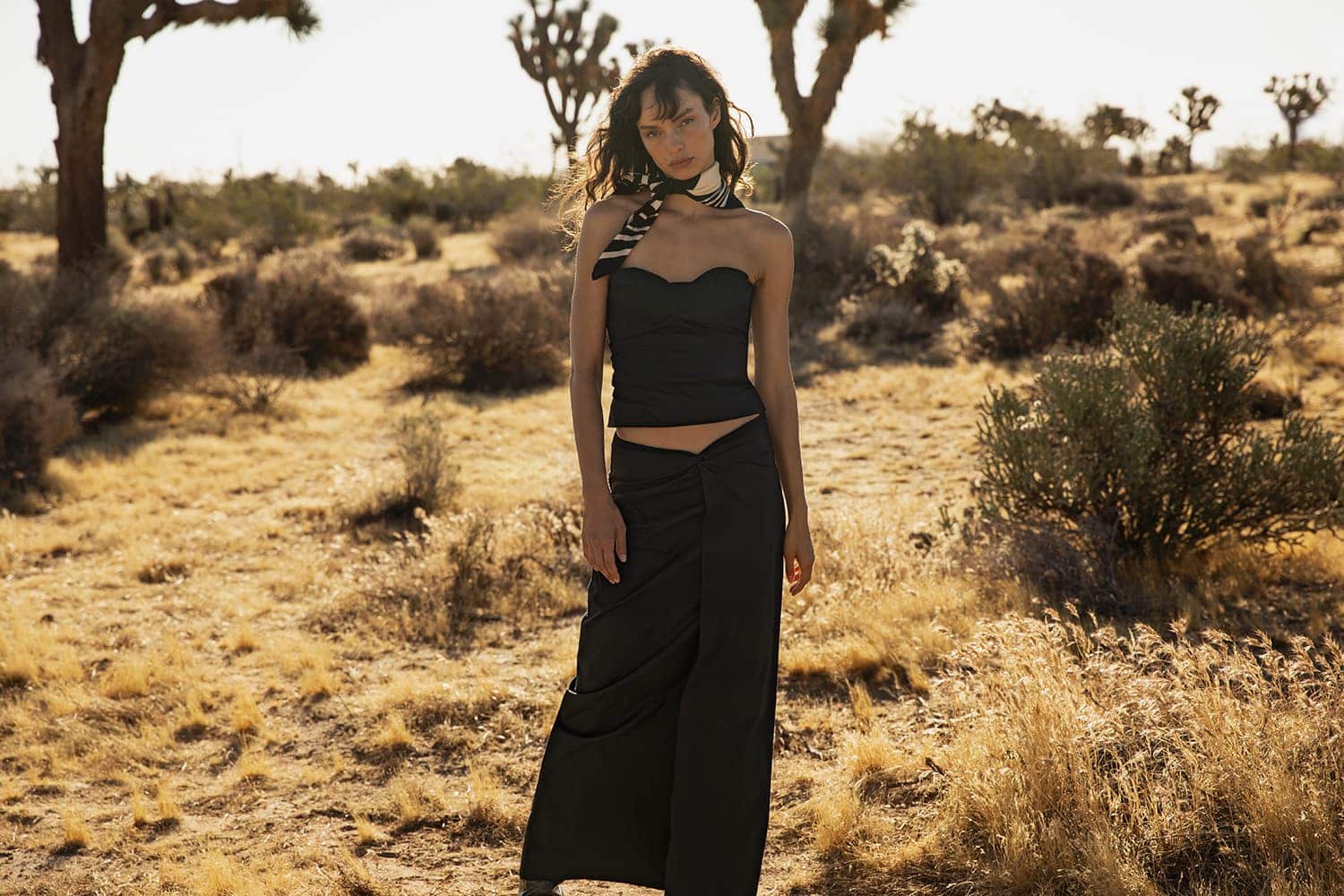 Woman poses in black top and skirt in a desert landscape with Joshua Trees and dry brush under sunny conditions, creating a fashion editorial.