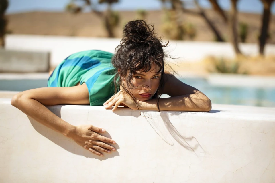 A woman with brown hair in a bun rests her arms on a white wall in front of a swimming pool and arid background in a portrait format.