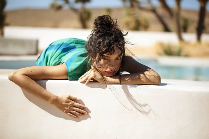 A woman with brown hair in a bun rests her arms on a white wall in front of a swimming pool and arid background in a portrait format.