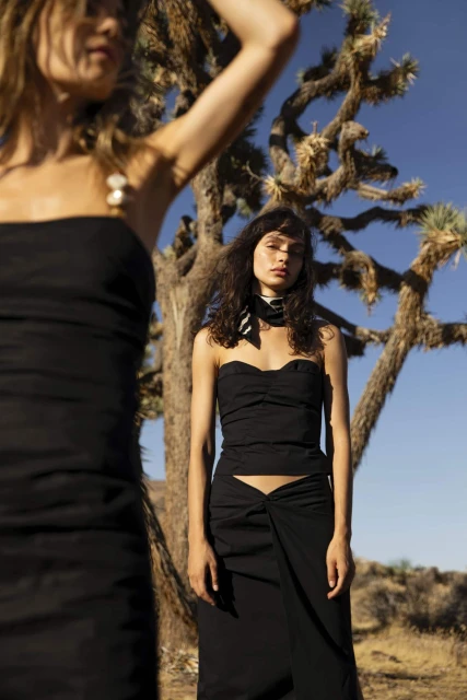Two women in black strapless tops and skirts pose in a desert setting with sparse vegetation and blue sky, fashion shoot, medium shot.