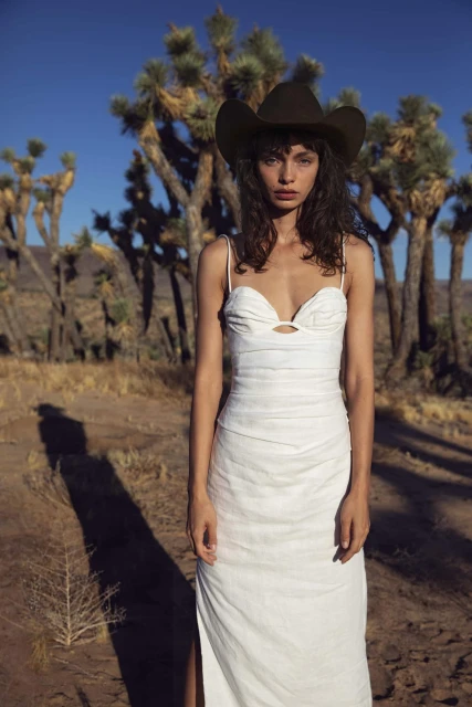 Brunette woman in an elegant white dress and a brown cowboy hat standing in the desert landscape with Joshua Trees on a sunny day.