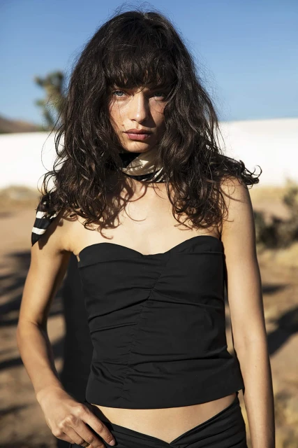 Brunette woman in a black strapless top, with a neck scarf, standing outdoors in a sunny desert environment. Fashion editorial portrait.