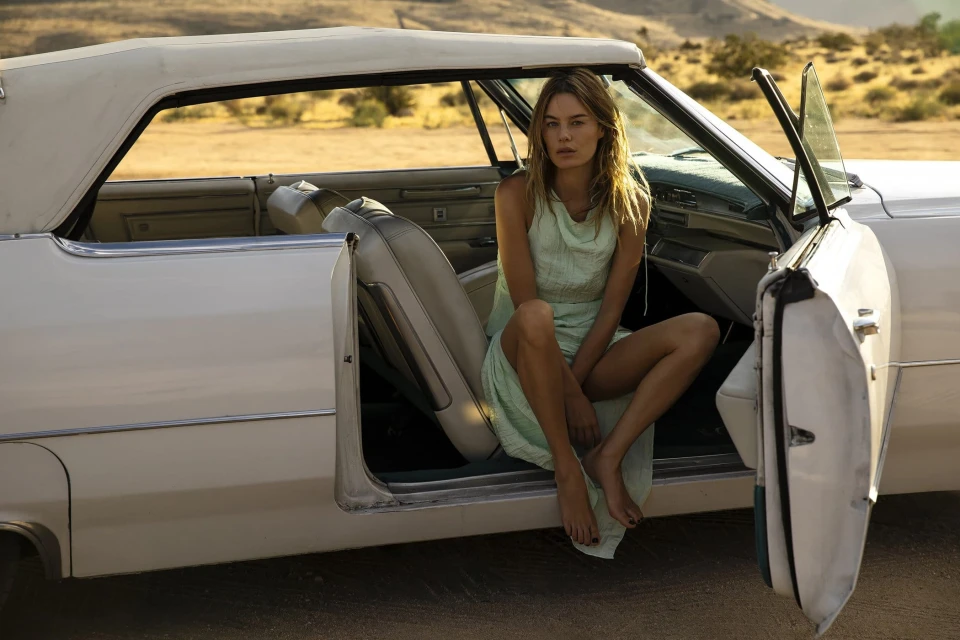 Young woman in a light green dress sitting inside a classic white convertible car in an outdoor desert setting during daylight.