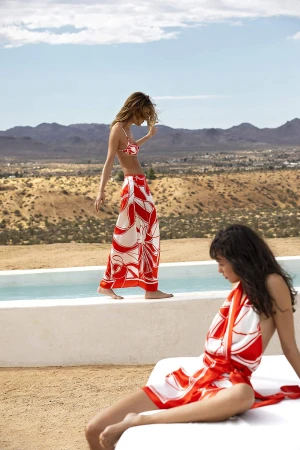 Two adult females model swimwear and sarongs with a red-and-white pattern by a pool in a desert landscape against a blue sky.