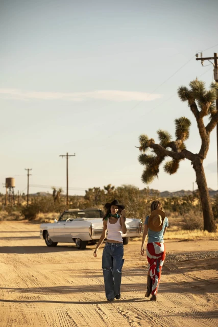 Two women walk on a dirt road in a desert landscape with a vintage car, sparse vegetation, and a clear sky in the background.