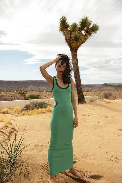 A slender woman in a green tank dress stands in a desert landscape with sparse vegetation and a single tall Joshua tree.
